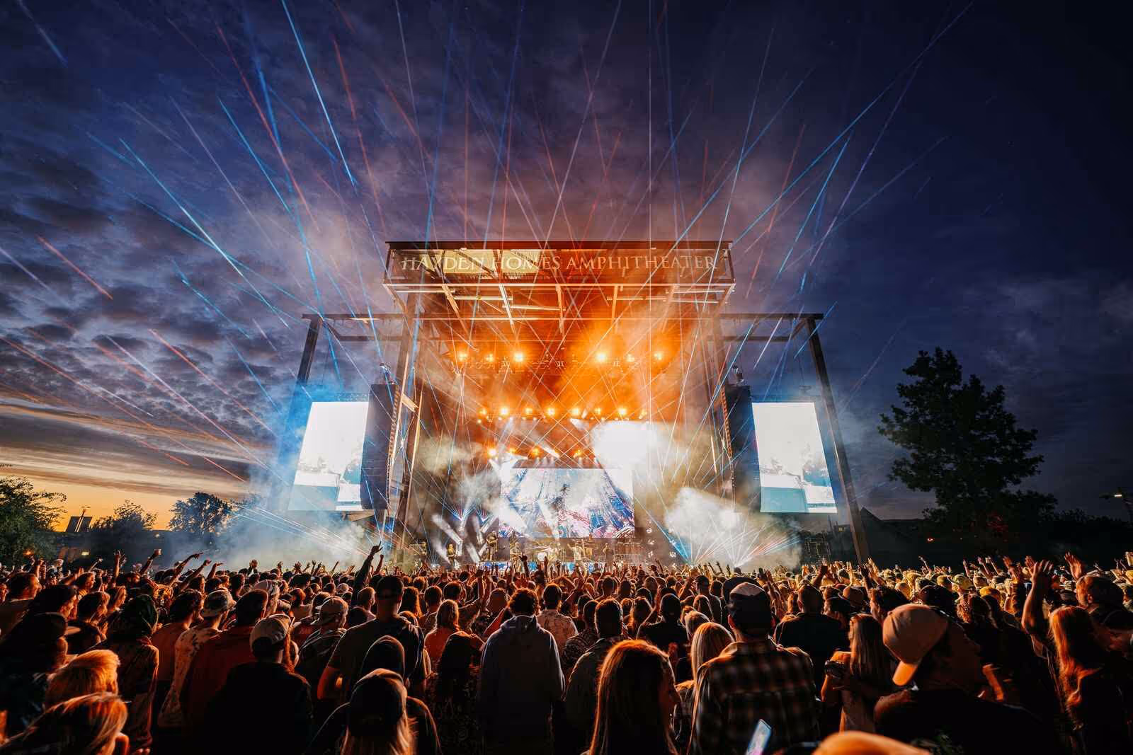 Crowd watching a brightly lit concert on an outdoor stage at dusk with light beams in the sky.