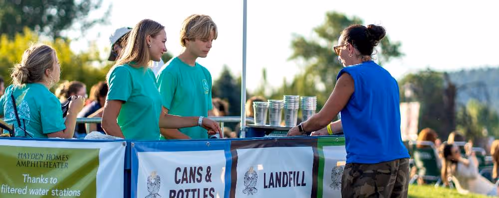 Volunteers in green shirts assist a woman recycling cans, bottles, and landfill waste outdoors.