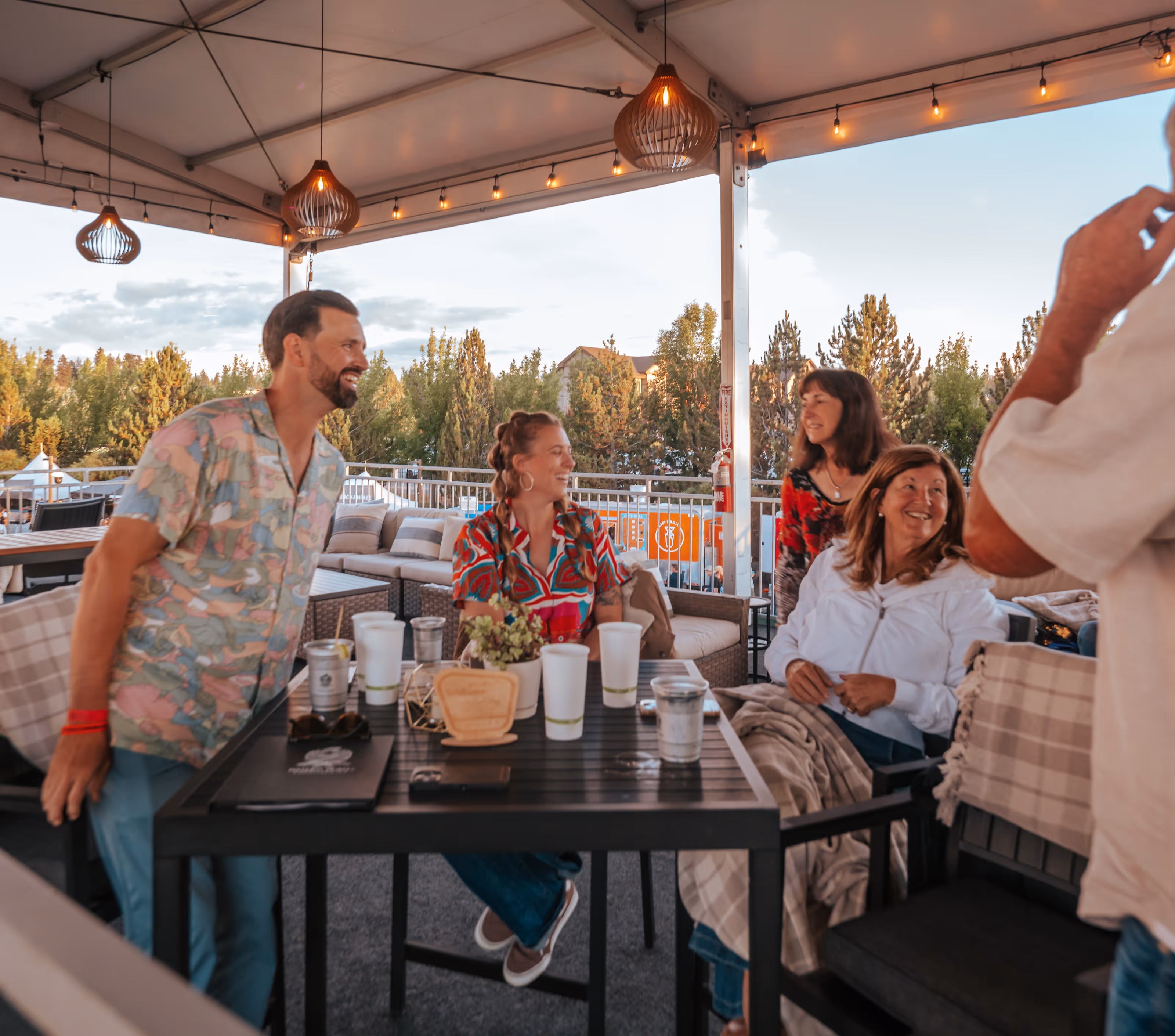 Four people smiling and talking at a table outdoors under a covered patio with string lights.