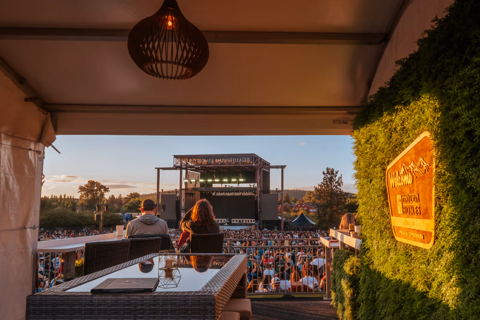 People seated on patio facing Hayden Homes Amphitheater stage during sunset with crowd below.