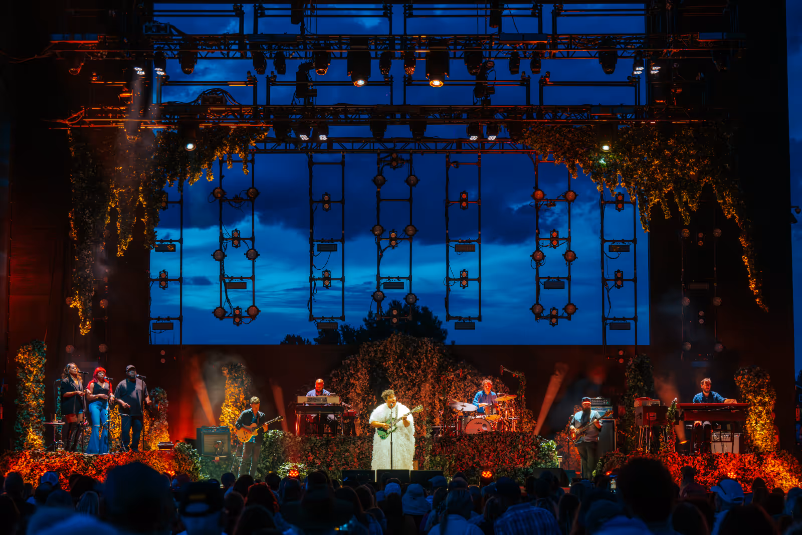 Band performing on outdoor stage decorated with greenery under a dusk sky, with audience watching.