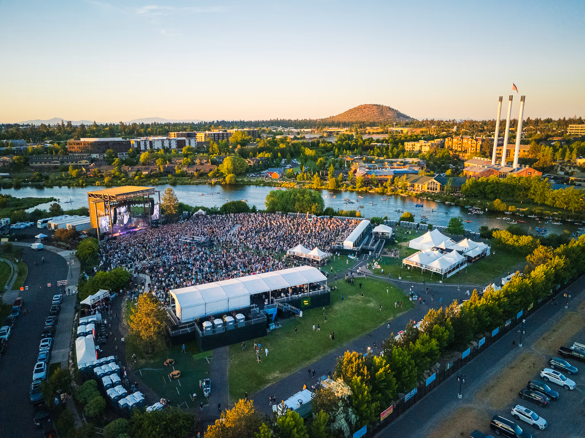 A large outdoor concert with a crowd near a river and three tall chimneys in the background at sunset.