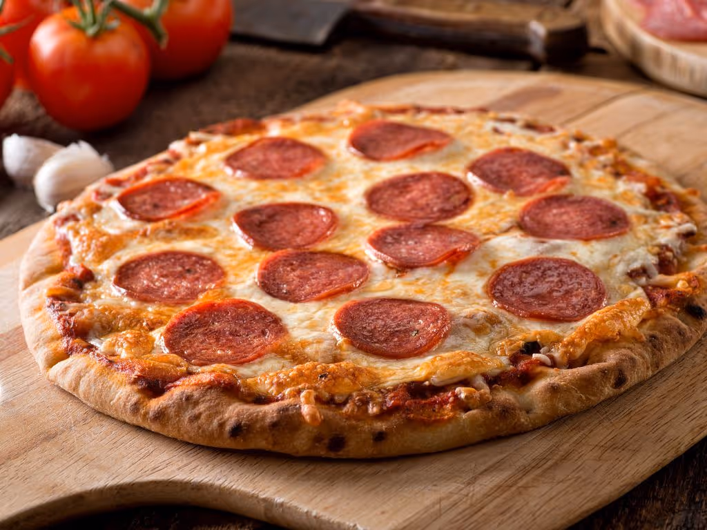 Pepperoni pizza with melted cheese on a wooden board, with tomatoes and garlic in the background.