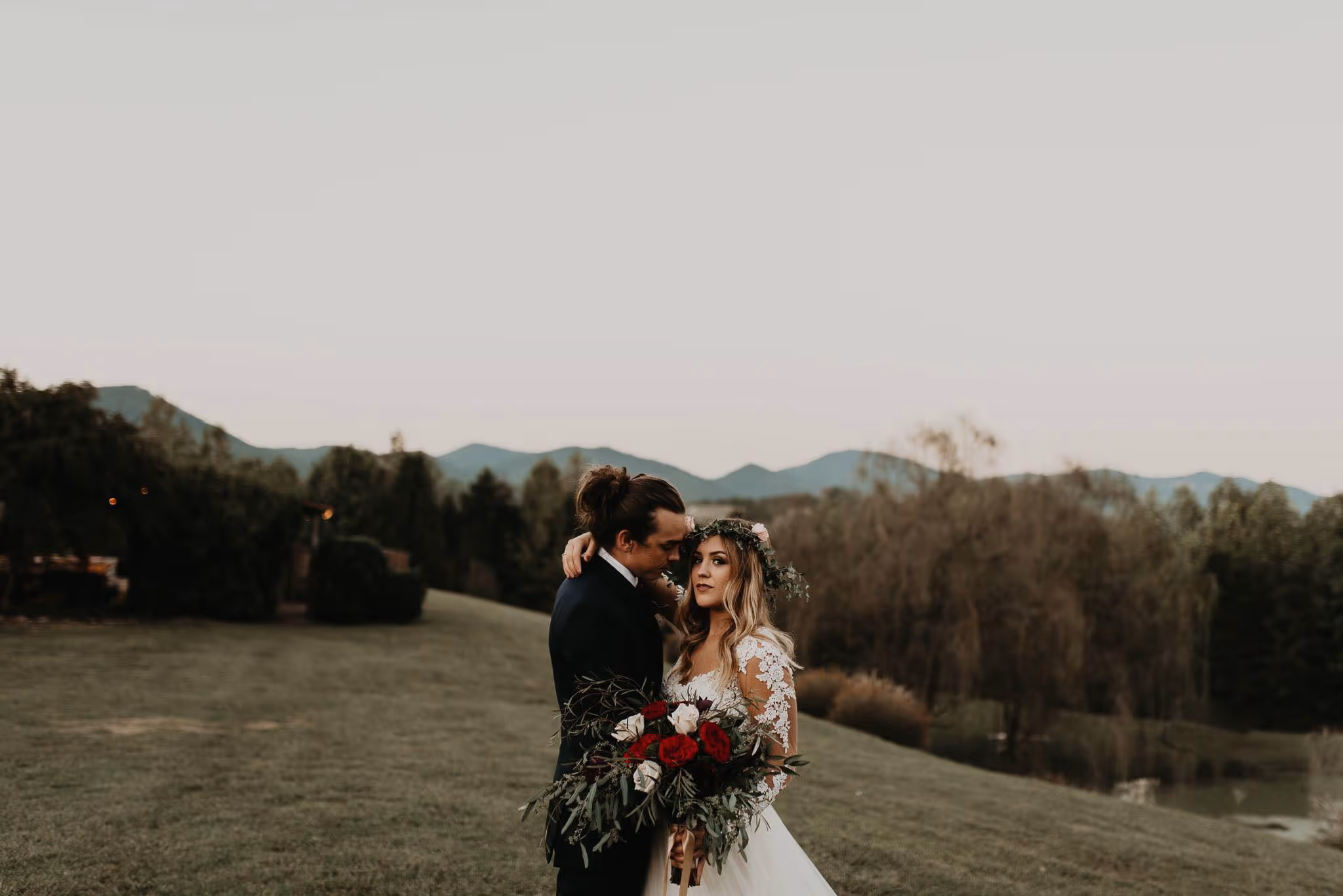 Bride in a white lace dress and flower crown holding a bouquet with red and white flowers, embraced by groom in a dark suit, standing outdoors with mountains and trees in the background.