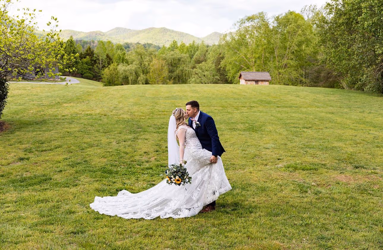Bride and groom kissing on a grassy field with trees and mountains in the background.