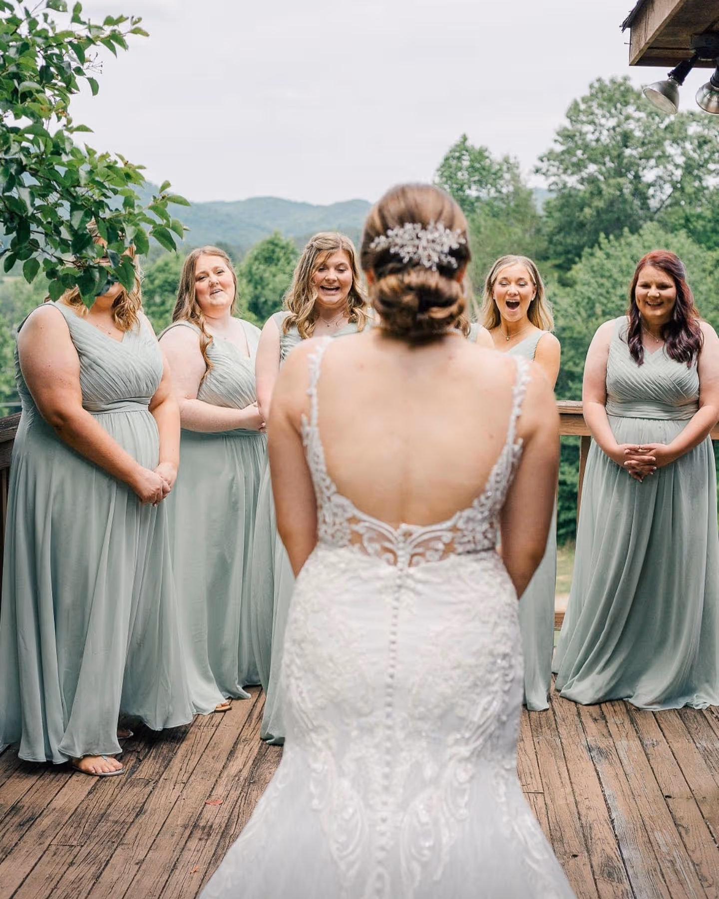 Bride in white lace gown with back to camera, facing five bridesmaids in sage green dresses on a wooden deck with trees and mountains in the background.
