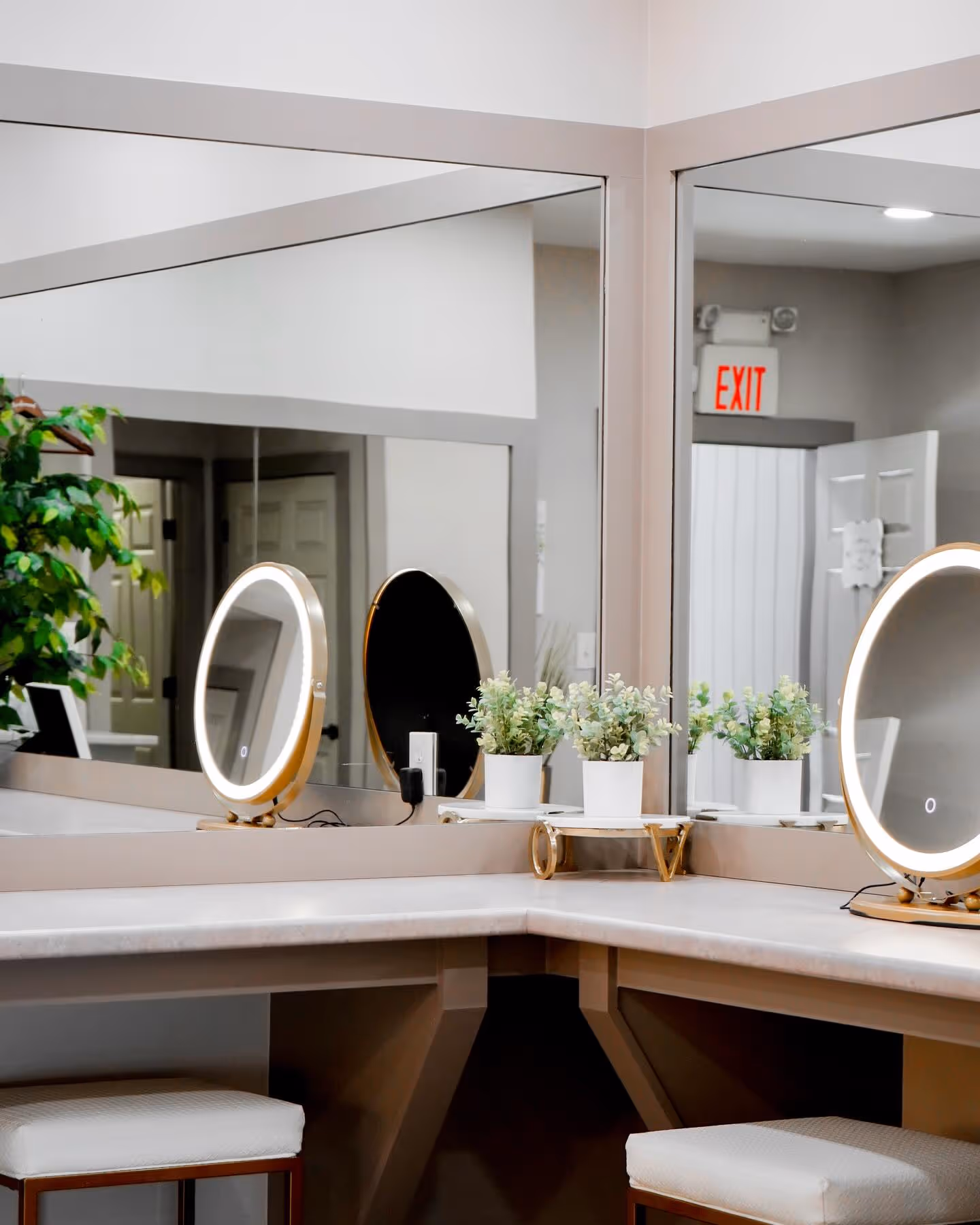 Vanity corner with two illuminated round mirrors, white cushioned stools, and potted plants in front of a large wall mirror.