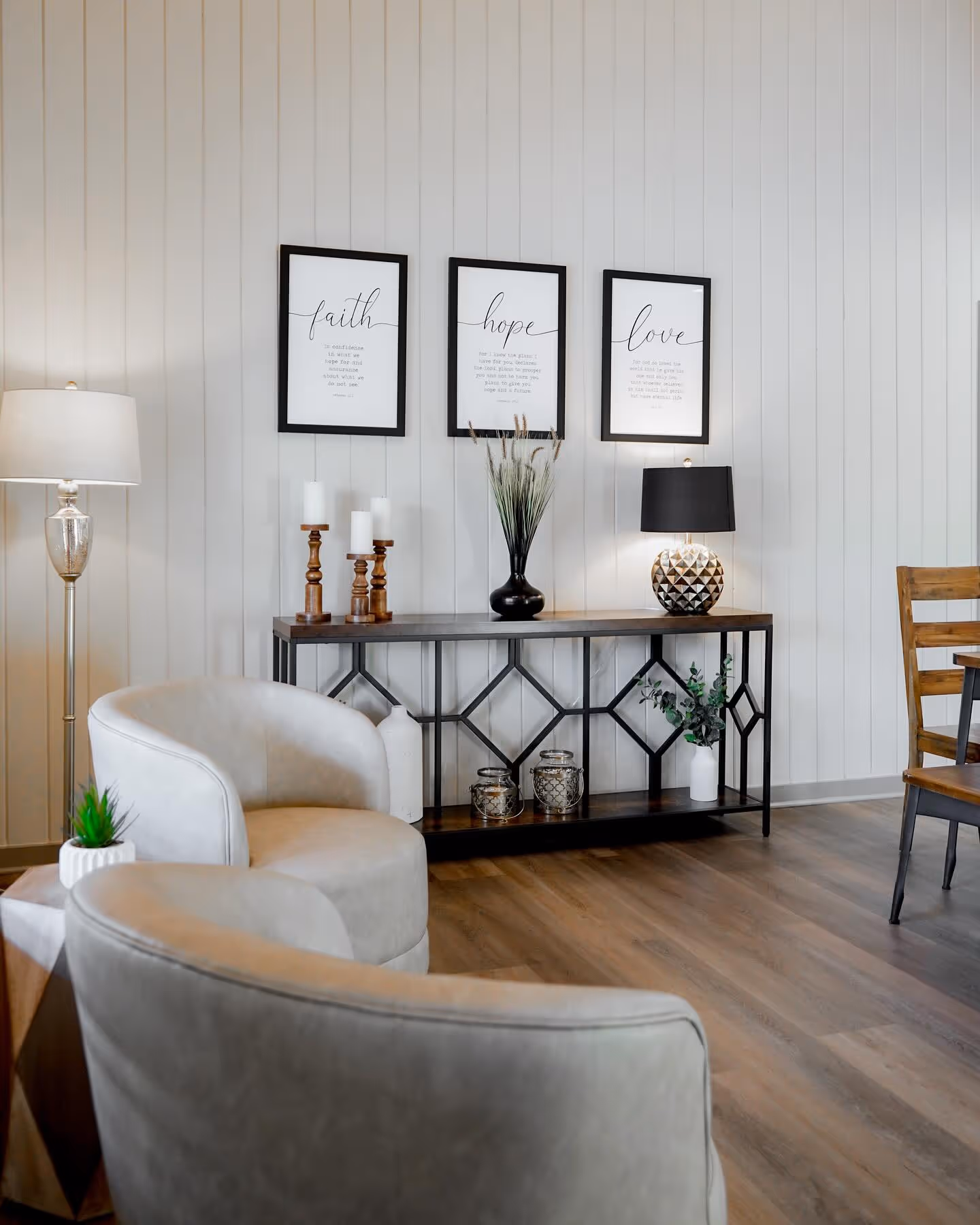 Modern living room with light gray armchairs, wooden floor, console table with candles, plants, lamps, and three framed inspirational quotes saying faith, hope, and love on a white paneled wall.