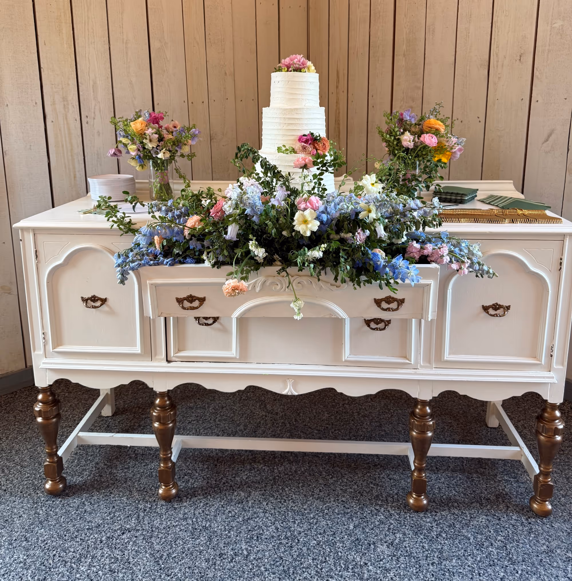 Three-tiered white wedding cake decorated with flowers on a vintage white sideboard adorned with blue and pink floral arrangements.
