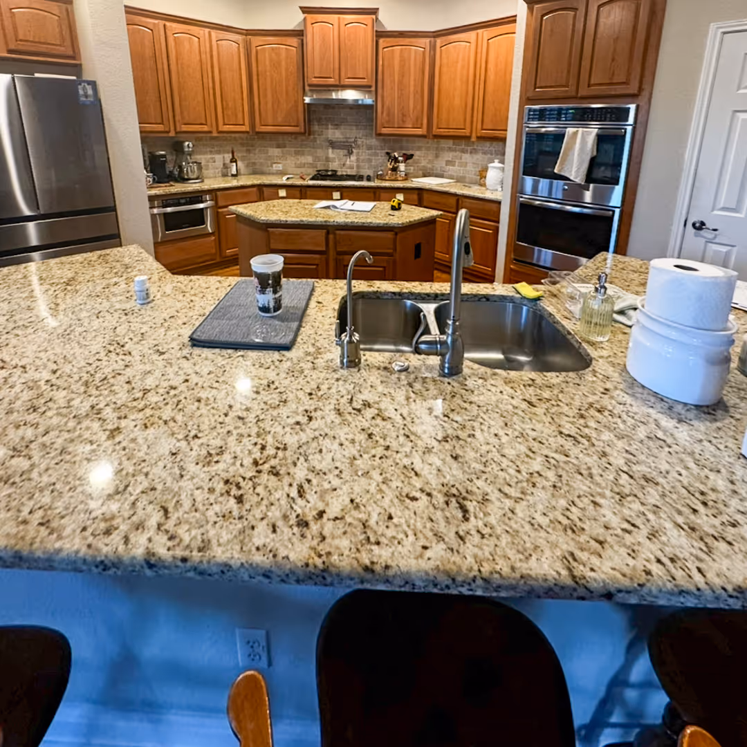 Modern kitchen before remodeling with granite countertops, wooden cabinets, stainless steel appliances, and a double sink.