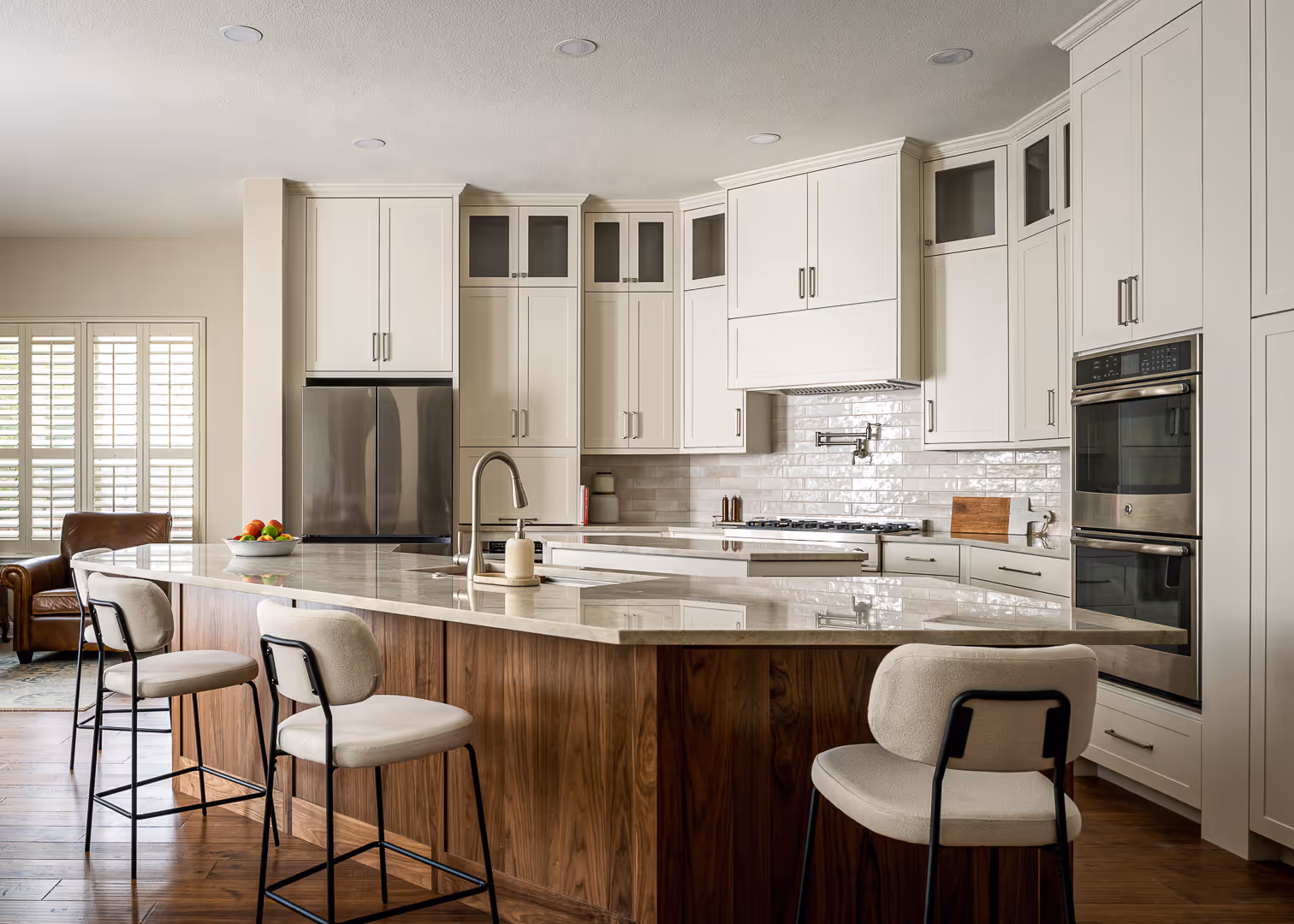 Modern kitchen remodel with white cabinets, a large marble island with wooden base, three cushioned bar stools, stainless steel refrigerator, and double wall ovens, in Cedar Park, TX.