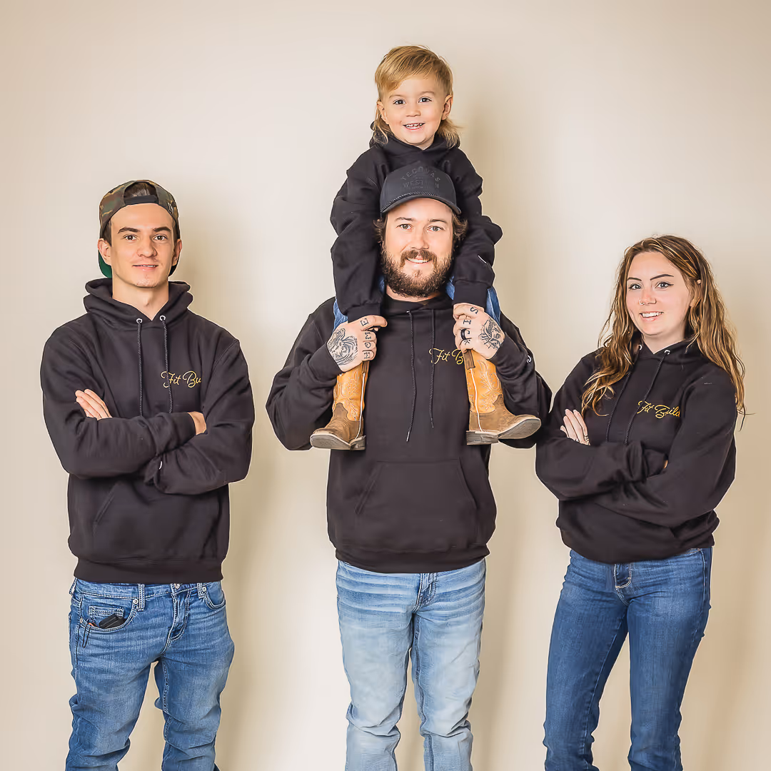Fit Build team wearing black hoodies and blue jeans with a young child sitting on the middle adult's shoulders, all smiling.