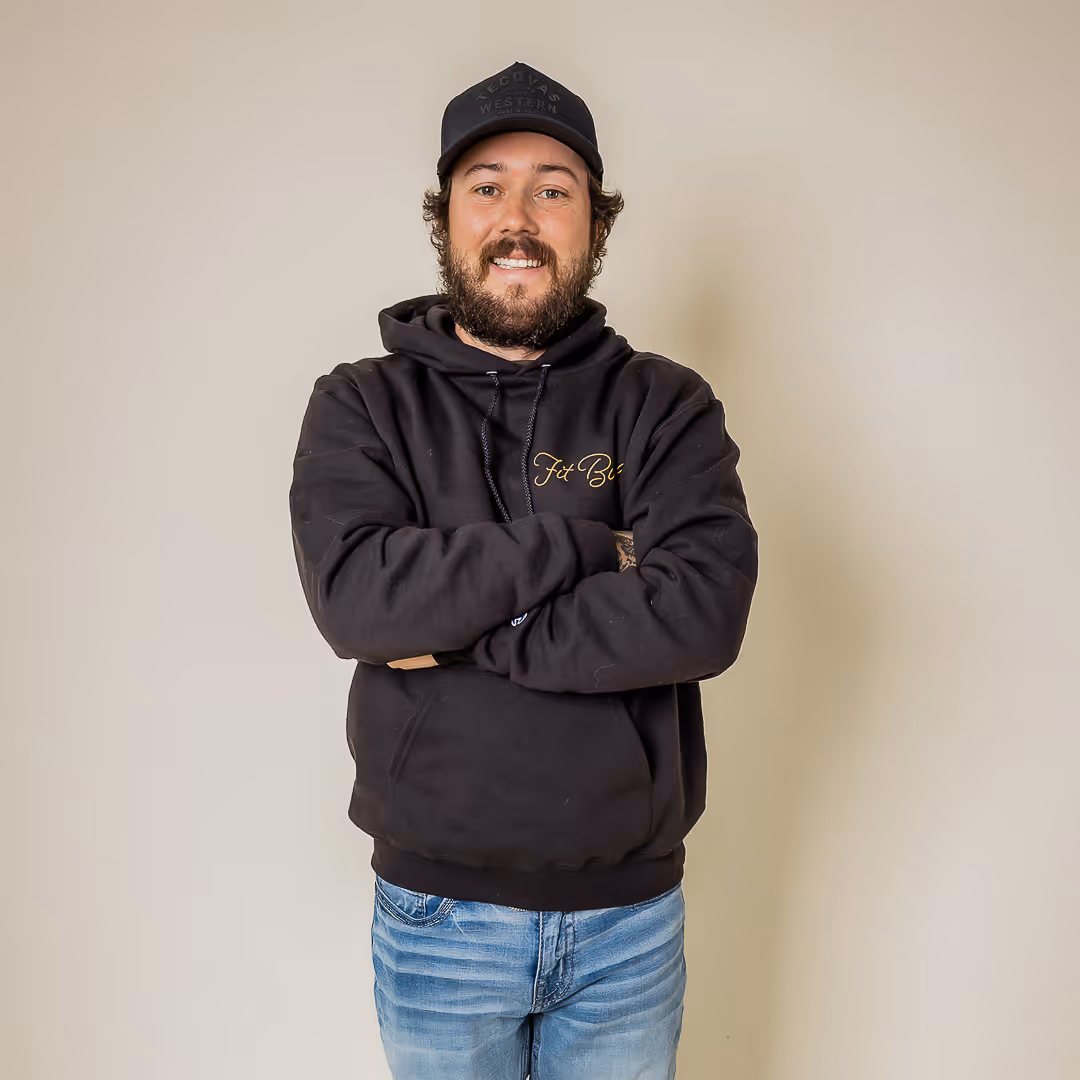 Smiling man with a beard wearing a black hoodie and black cap standing with arms crossed against a plain beige background.