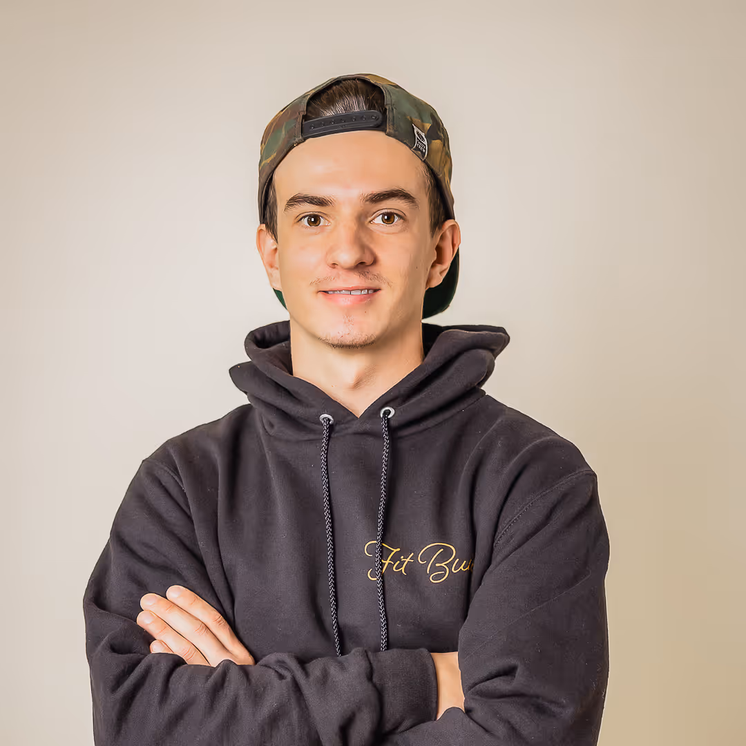 Young man wearing a black hoodie and camo baseball cap with arms crossed, smiling at the camera.