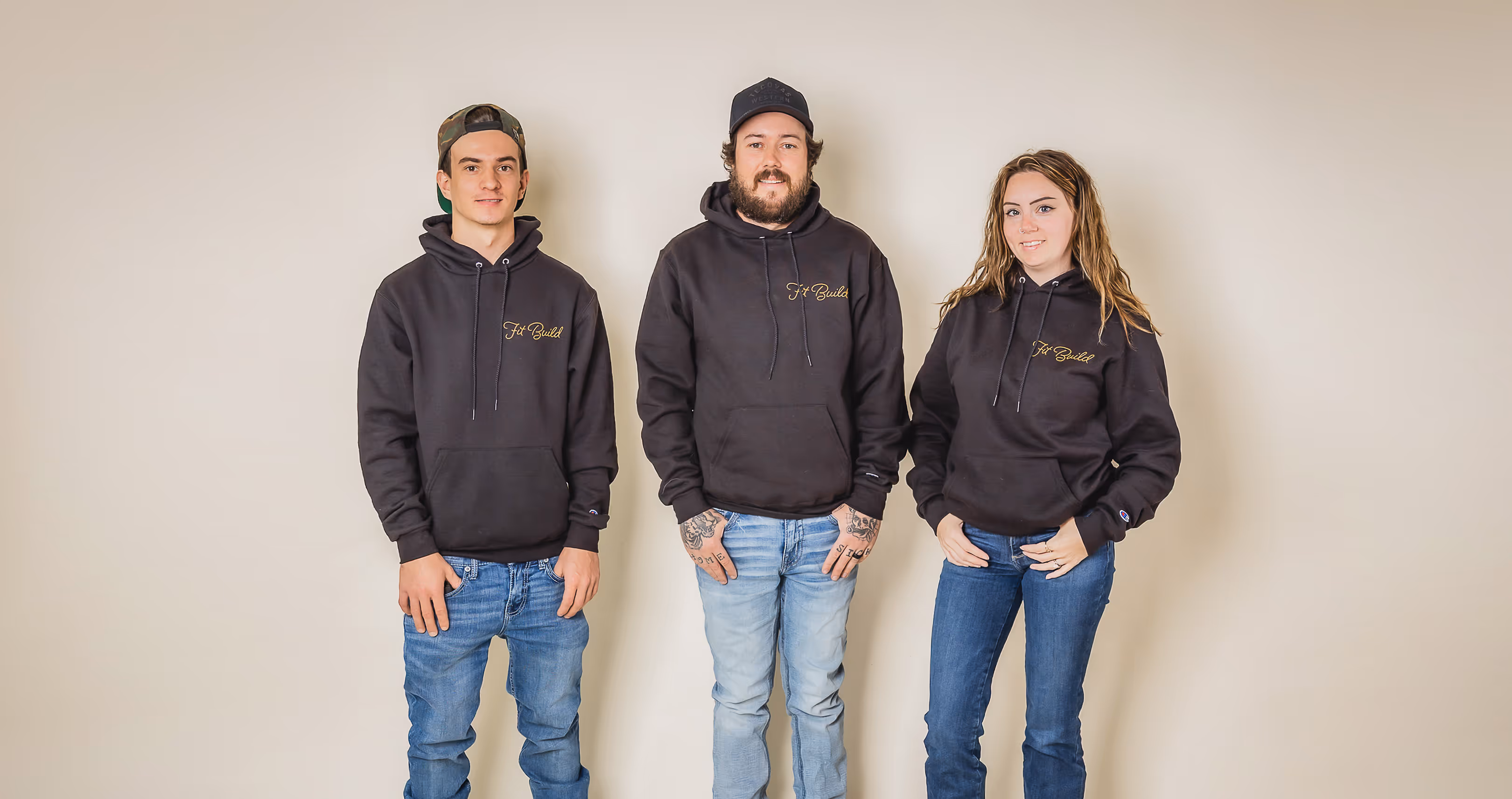Fit Build Team standing in a row wearing matching black hoodies with yellow text and blue jeans against a plain beige background.