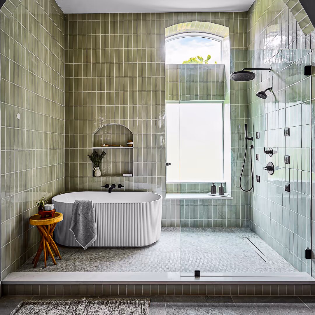 Modern bathroom with olive green tiled walls, a white freestanding tub, glass-enclosed shower with rainfall showerhead, and a small wooden side table with plants and toiletries.
