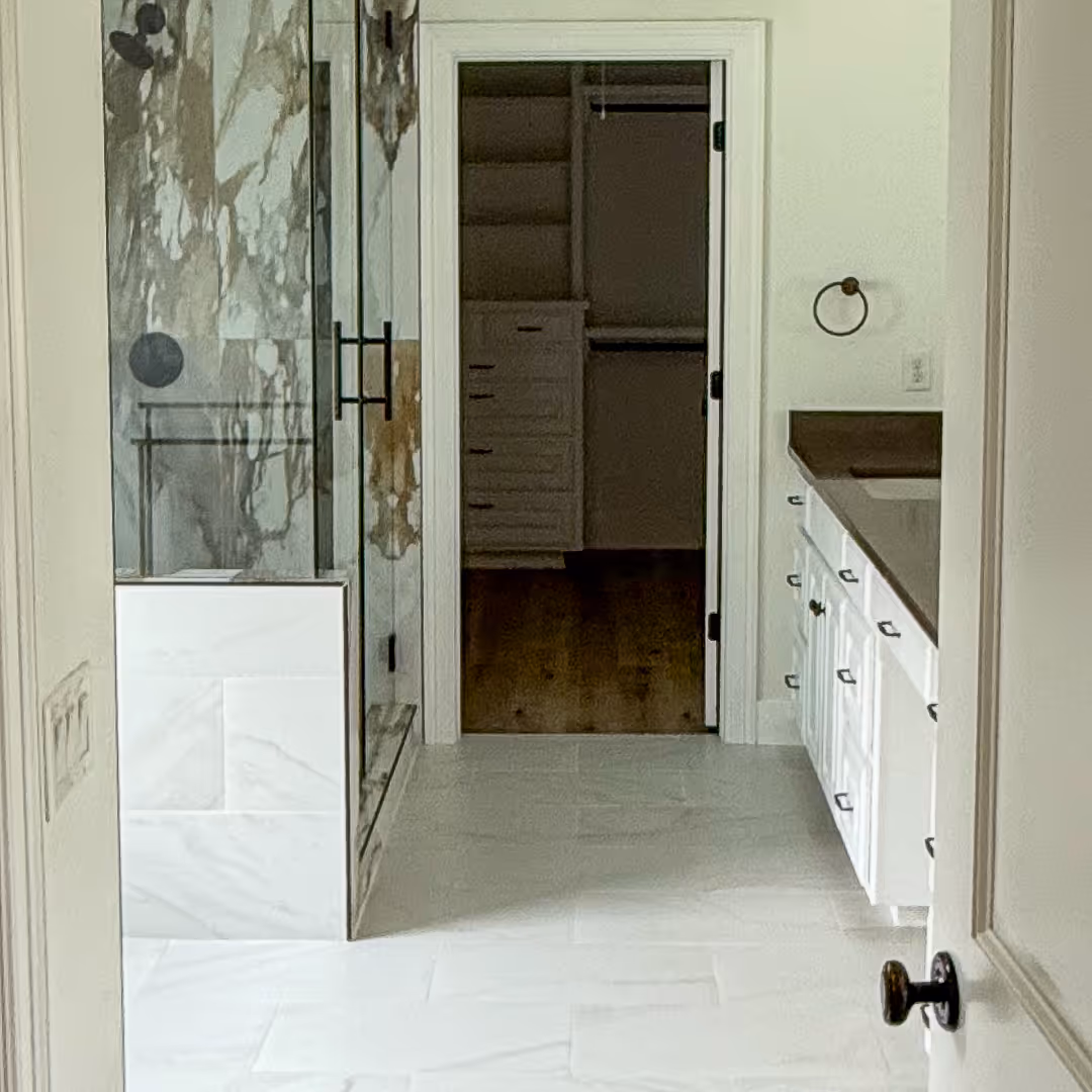 Before remodel - bathroom with white tiles, glass shower enclosure with marble walls, white vanity with dark countertop, and open walk-in closet in the background, Tarrytown, Austin.