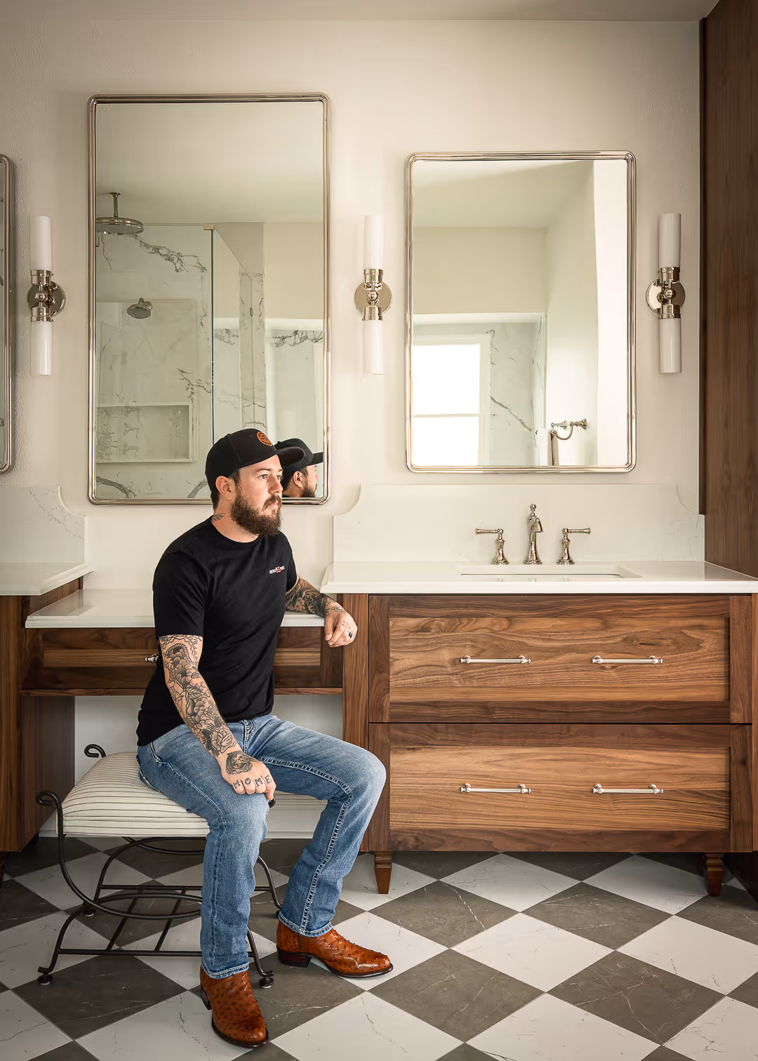Contractor wearing a black t-shirt, jeans, and brown boots sitting on a striped stool in a bathroom with wood vanity, marble countertop, and two mirrors.