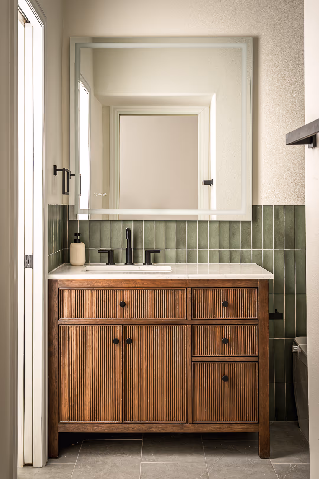 Bathroom vanity with vertical wood slat cabinet doors, white countertop, black faucet, green vertical tile backsplash, and a large square mirror, in Austin.
