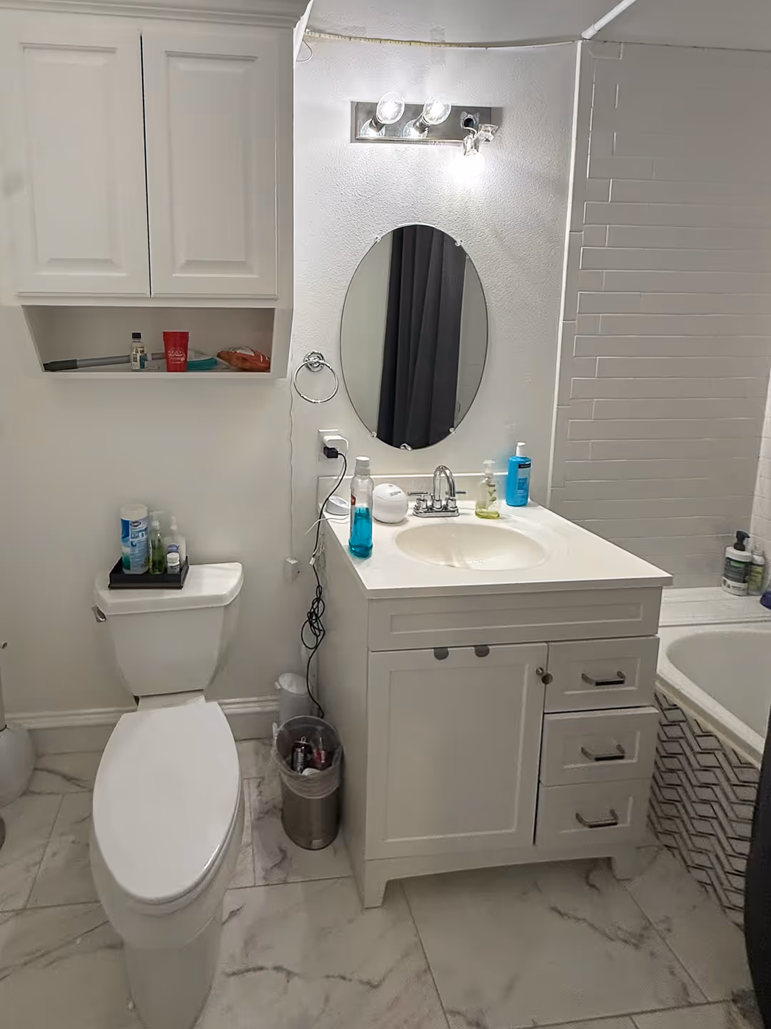 Compact Austin bathroom with white toilet, white vanity with oval mirror, shelves with toiletries, and a bathtub with white tile surround, before remodel.