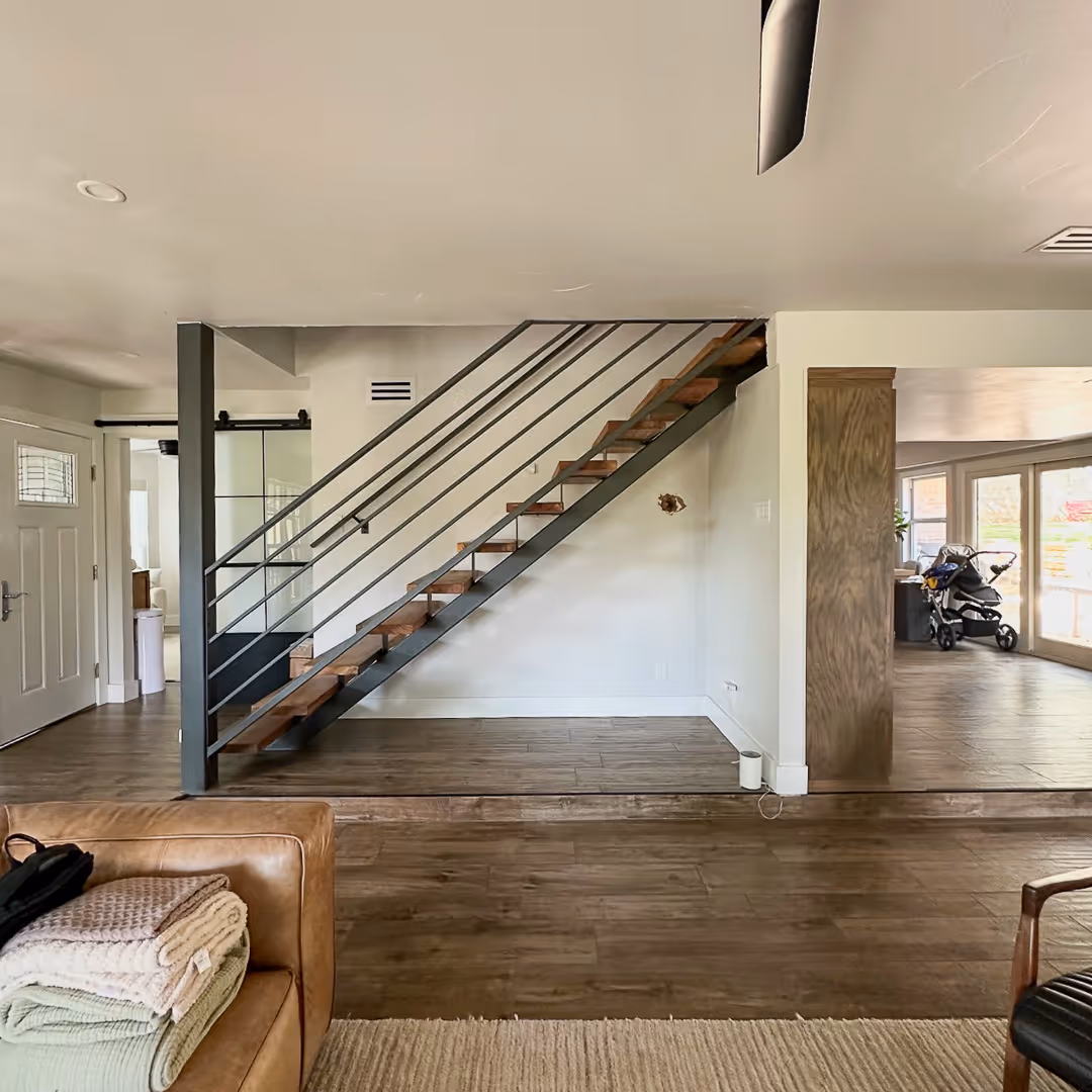 Before interior with wooden stairs featuring metal railings, brown leather sofa with folded blankets, and a baby stroller near large windows.