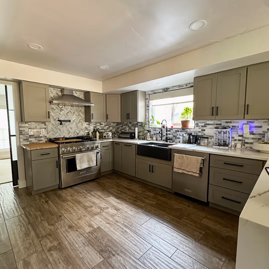 Modern kitchen with gray cabinets, stainless steel stove, black farmhouse sink, mosaic tile backsplash, and wood-look floor.