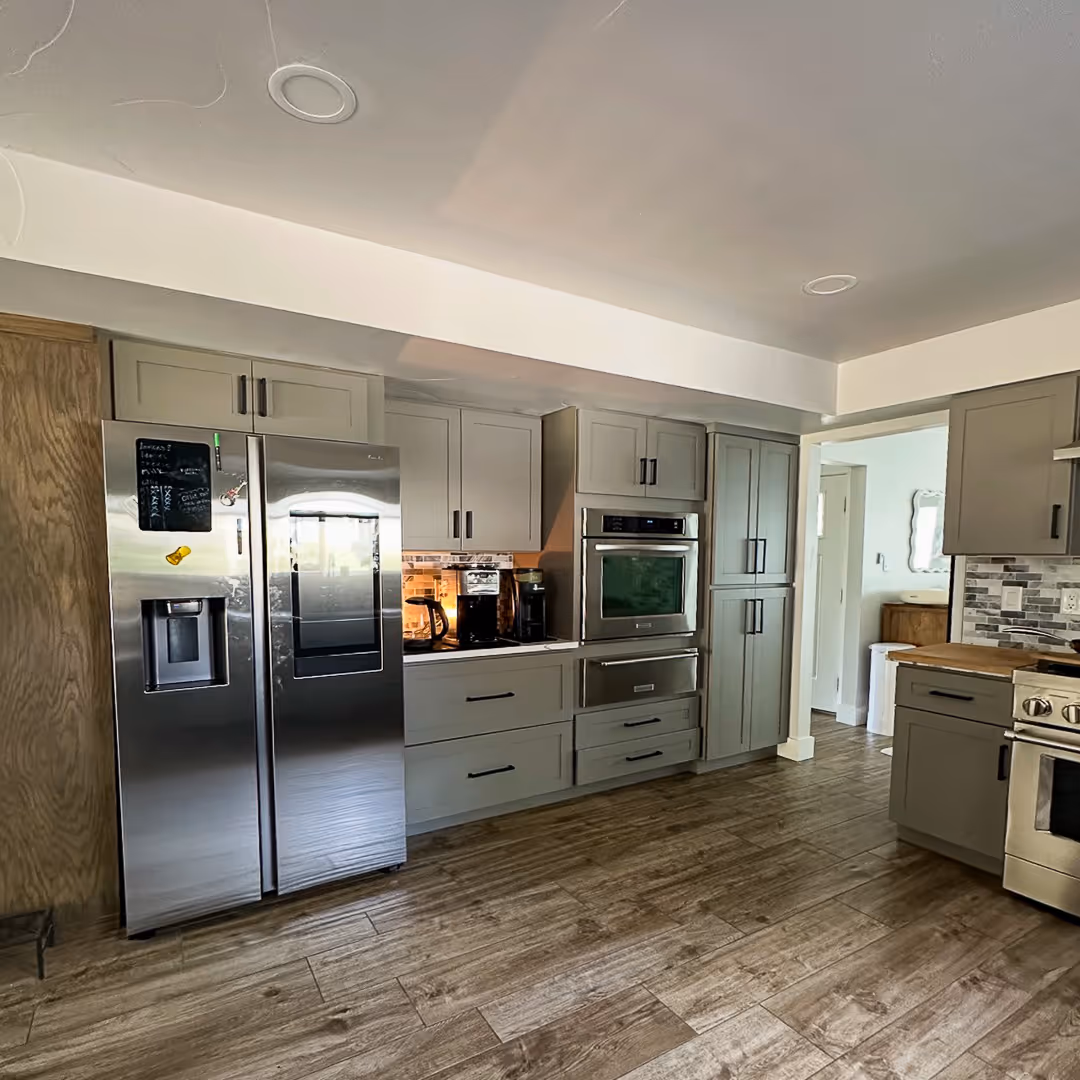 Modern kitchen with gray cabinets, stainless steel refrigerator, built-in oven, and wooden flooring.