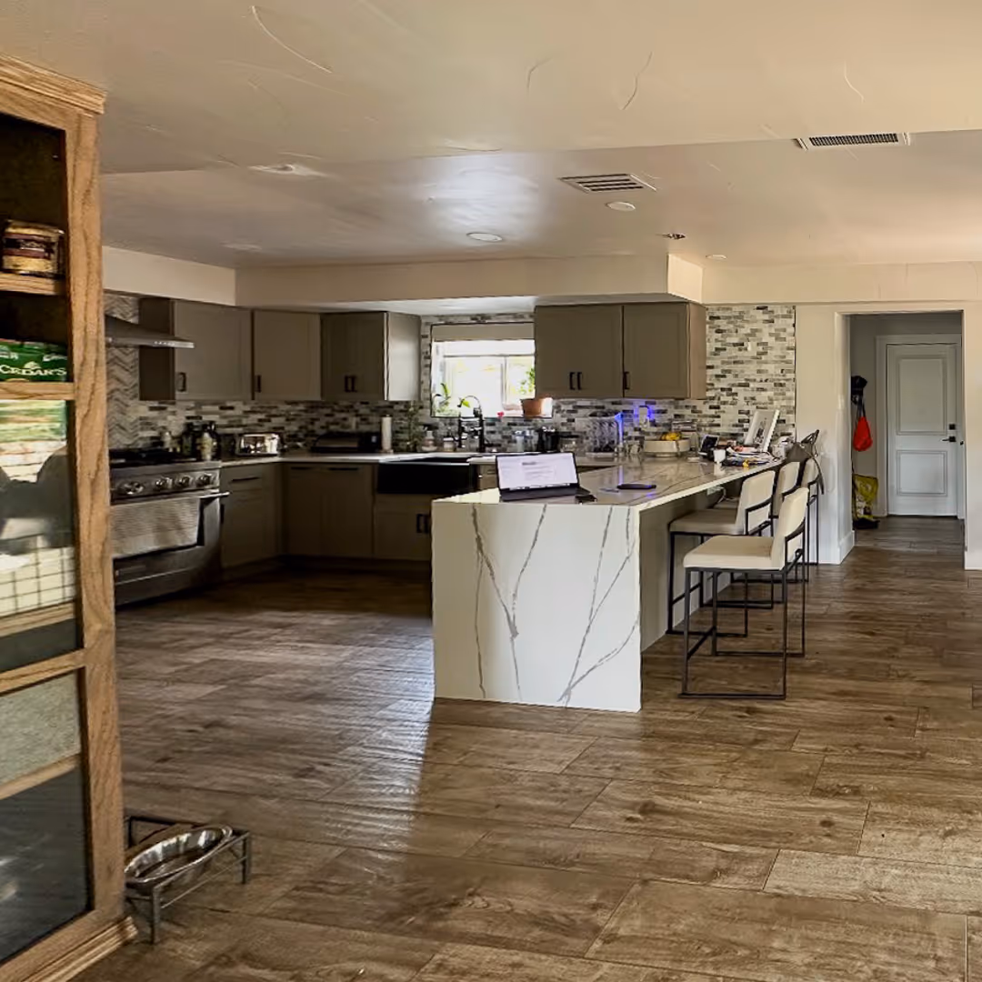 Modern kitchen with wooden floor, gray cabinets, marble island with three stools, and a window above the sink.