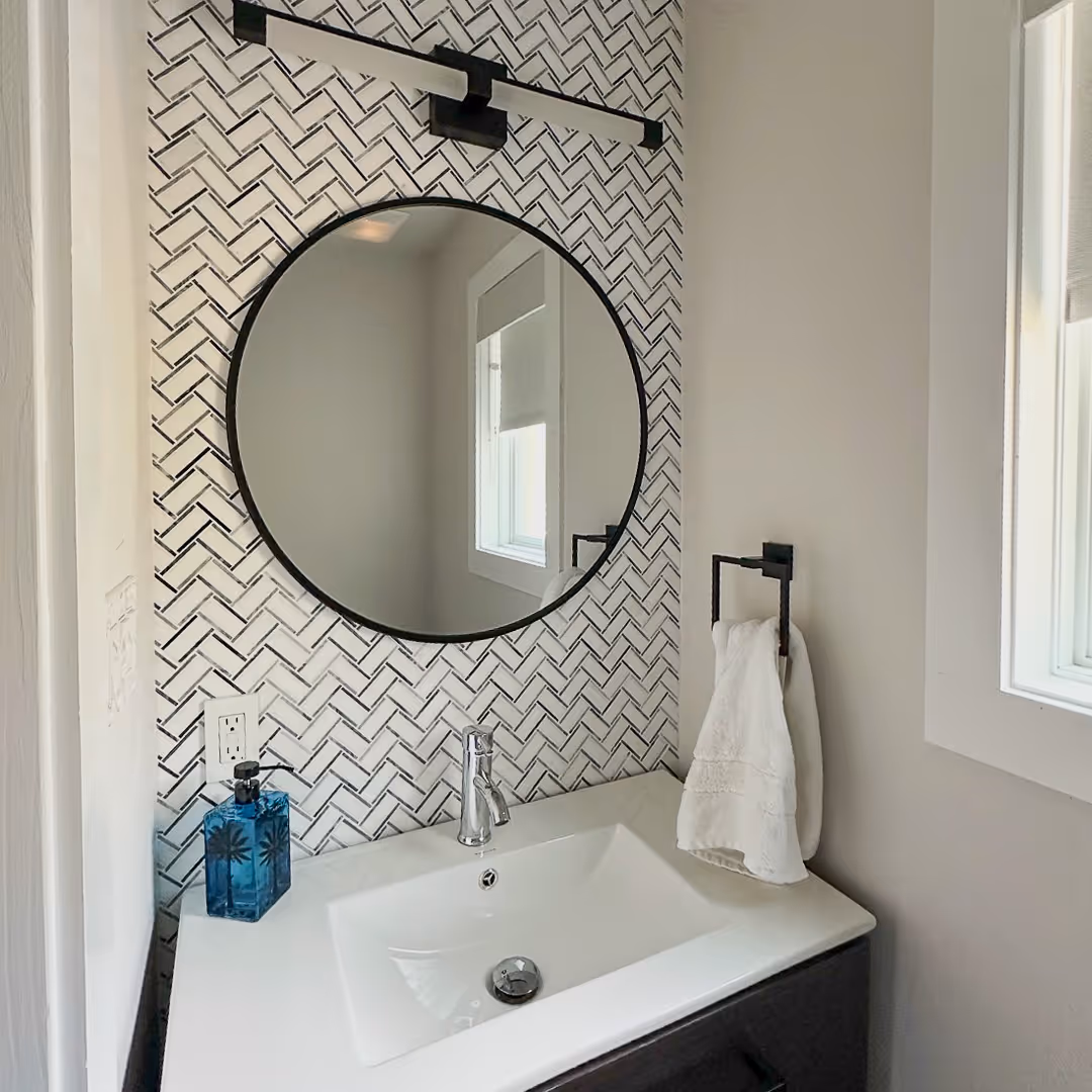 Modern bathroom vanity with white rectangular sink, round black-framed mirror, black towel ring holding white towel, and patterned tile backsplash.