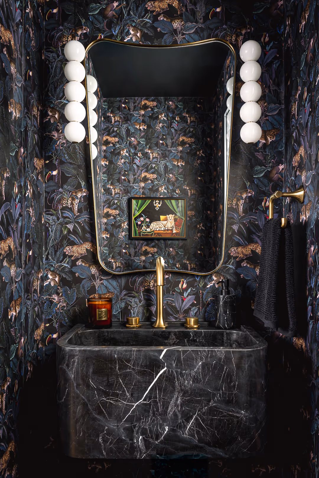 Bathroom  remodel with dark jungle-themed wallpaper featuring leaves, leopards, and monkeys, a large gold-framed mirror, black marble sink, and brass fixtures with white globe sconces and a black towel.