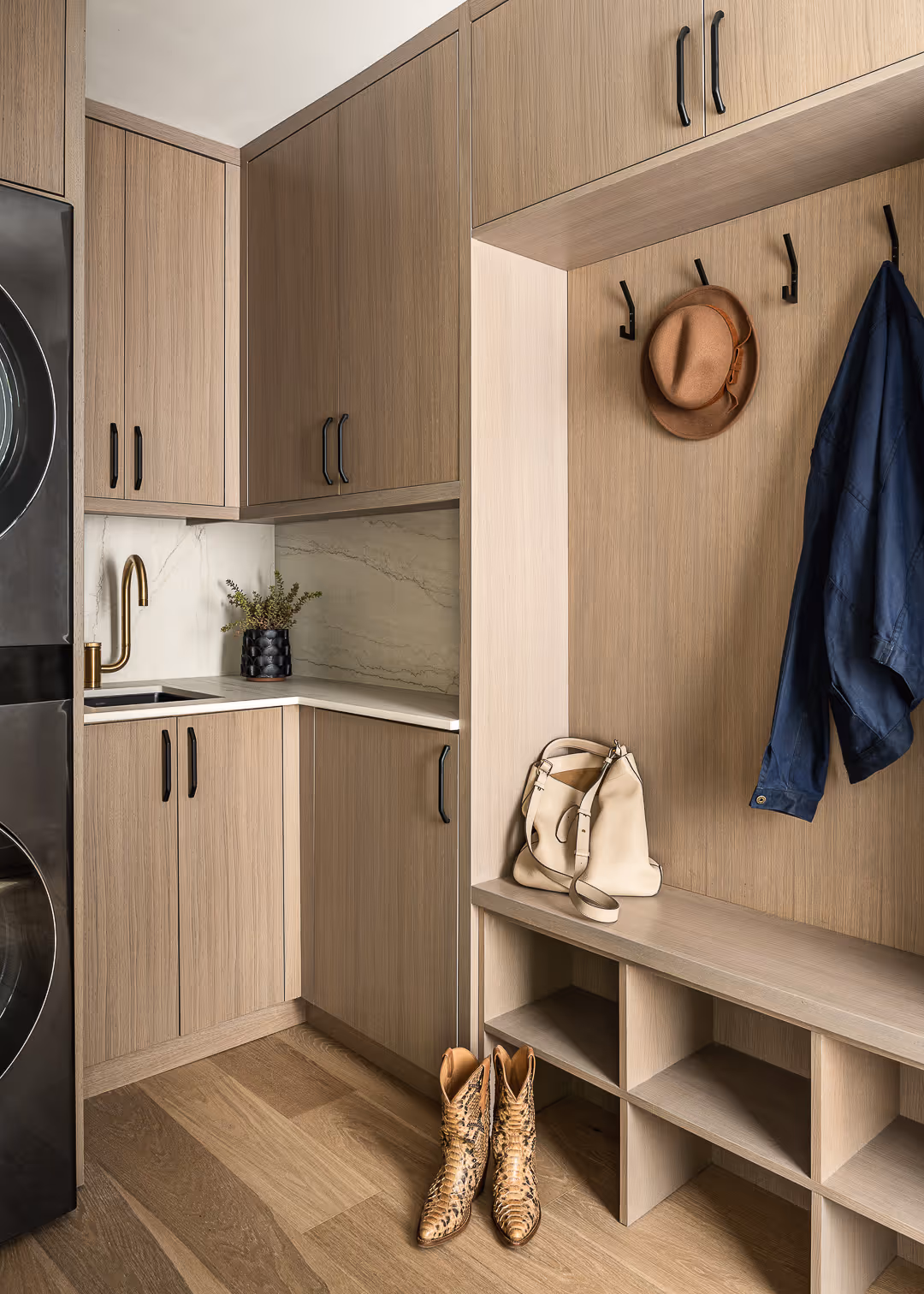 Minimalist laundry room with light wood cabinets, a stacked washer and dryer, beige handbag on bench, cowboy boots on floor, a brown hat and navy jacket hanging on black hooks, and a marble backsplash with gold faucet.