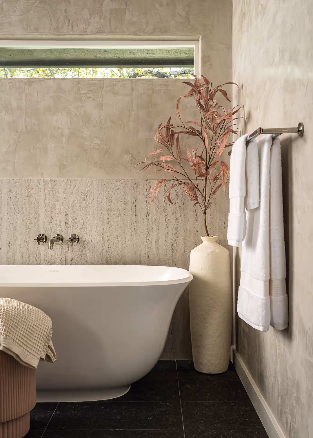 Modern bathroom remodel with a white freestanding bathtub, textured beige walls, a tall beige vase with reddish leaves, and white towels hanging on a metal towel rack.
