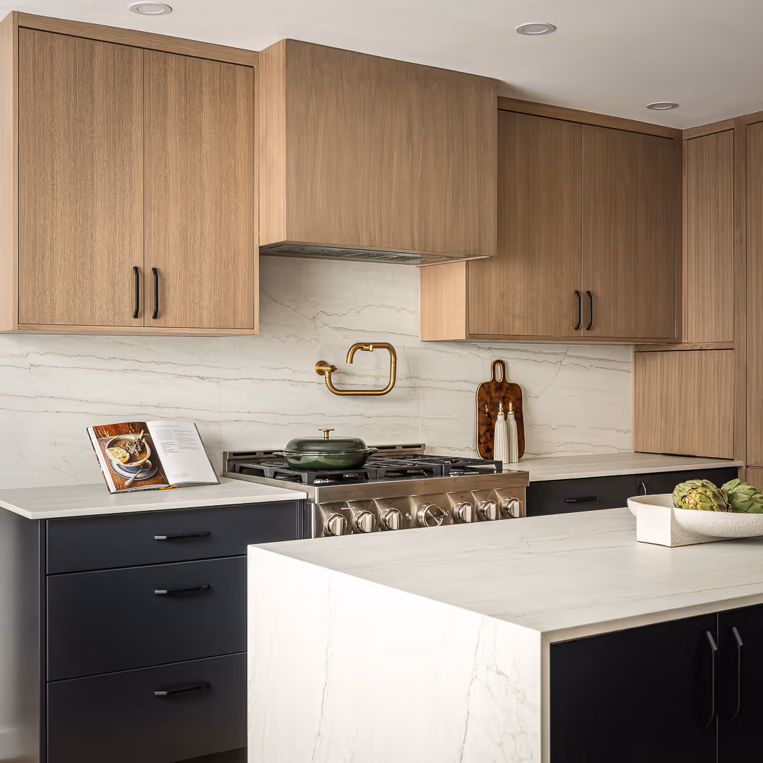 Modern remodeled kitchen with wooden upper cabinets, black lower drawers, white marble countertops, a stainless steel gas stove with a green pot, and a gold wall-mounted faucet, in Austin.