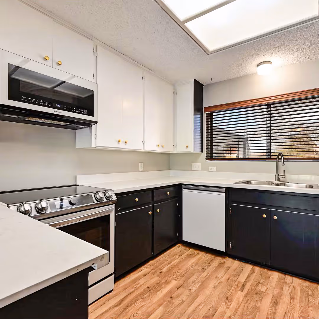 Before the remodel kitchen with white upper cabinets, black lower cabinets, stainless steel stove and microwave, white countertops, and wood flooring.