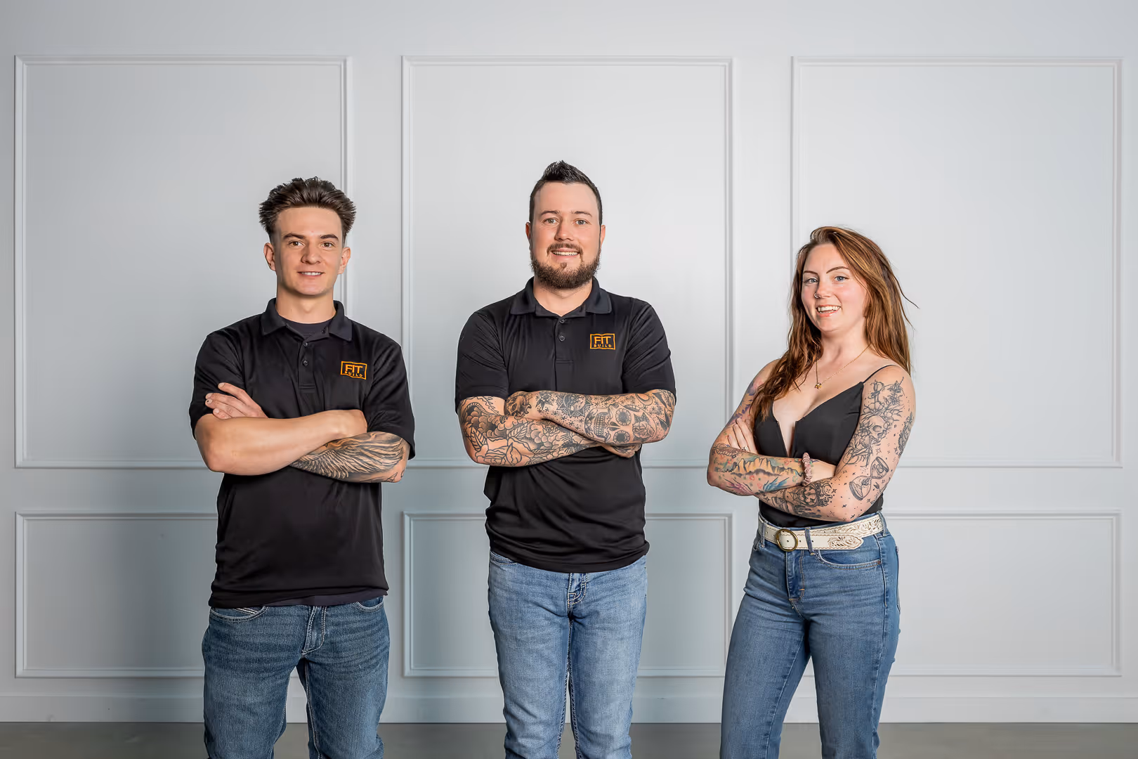 Fit Build team standing against a beige background wearing matching black hoodies and blue jeans, smiling at the camera.
