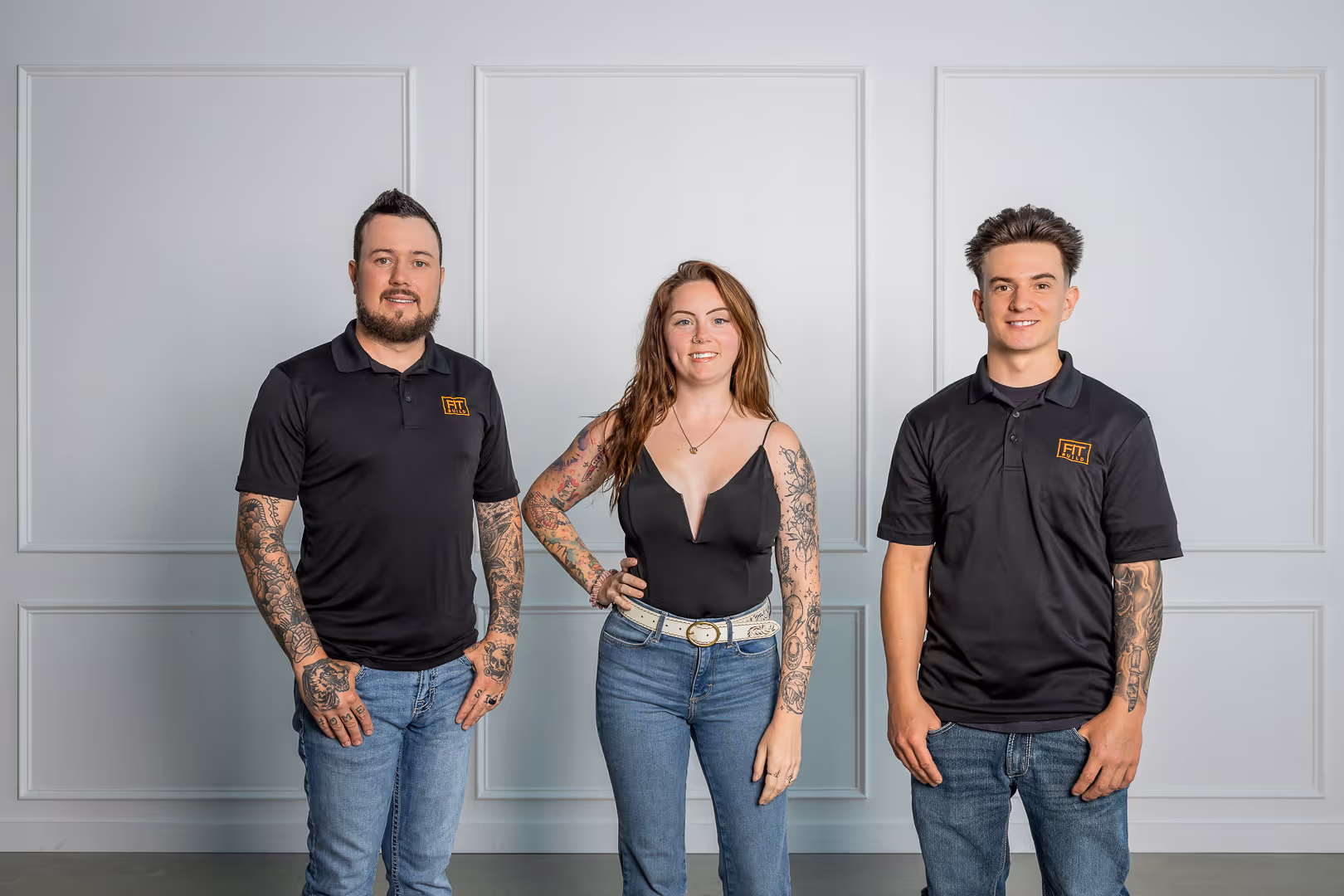Fit Build Team standing in a row wearing matching black hoodies with yellow text and blue jeans against a plain beige background.