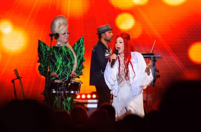 The B-52s in wild green and white flowing outfits with bright red lights in the background