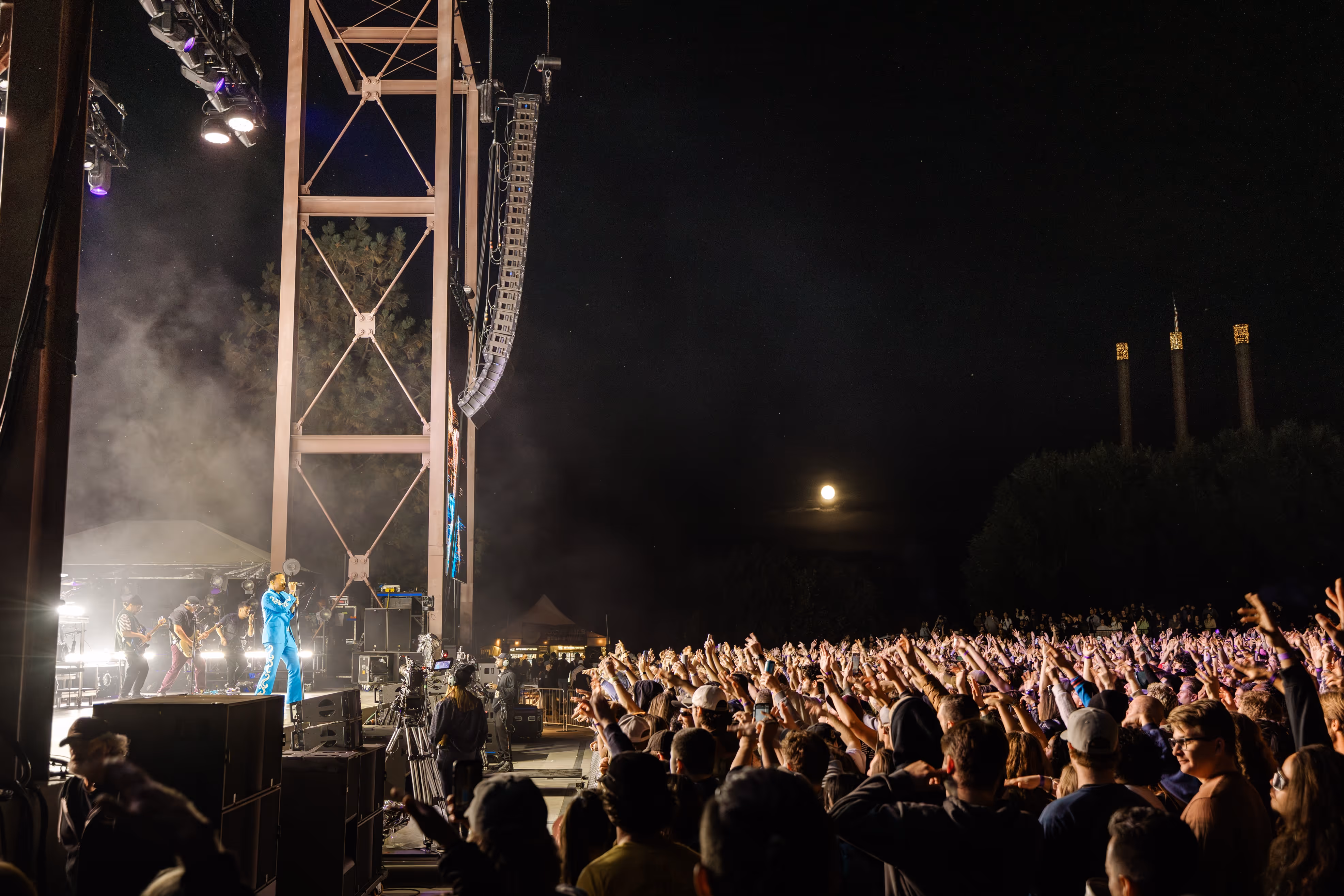 Anderson .Paak in a bright blue suit singing to a cheering crowd with a full moon rising in the background at Hayden Homes Amphitheater in Bend, Oregon