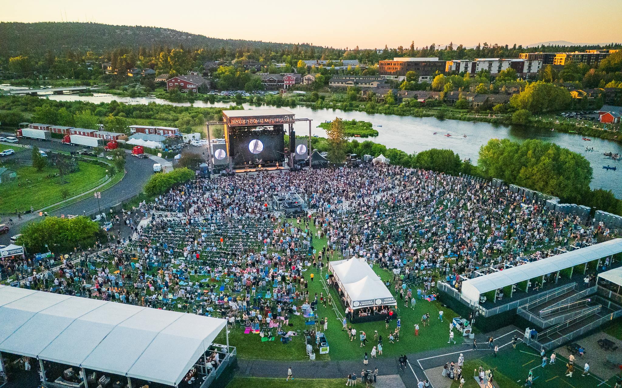Aerial view of Hayden Homes Amphitheater in Bend, Oregon from back of venue with the rail cars, full view of the stage and the Deschutes River in frame.