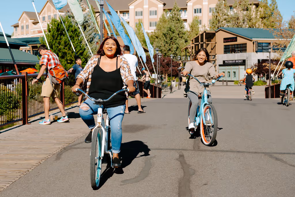 Gal with brown and pink hair riding her bike over the flag bridge in the Old Mill District in Bend, Oregon laughing with her friend on a baby blue bike beside her.