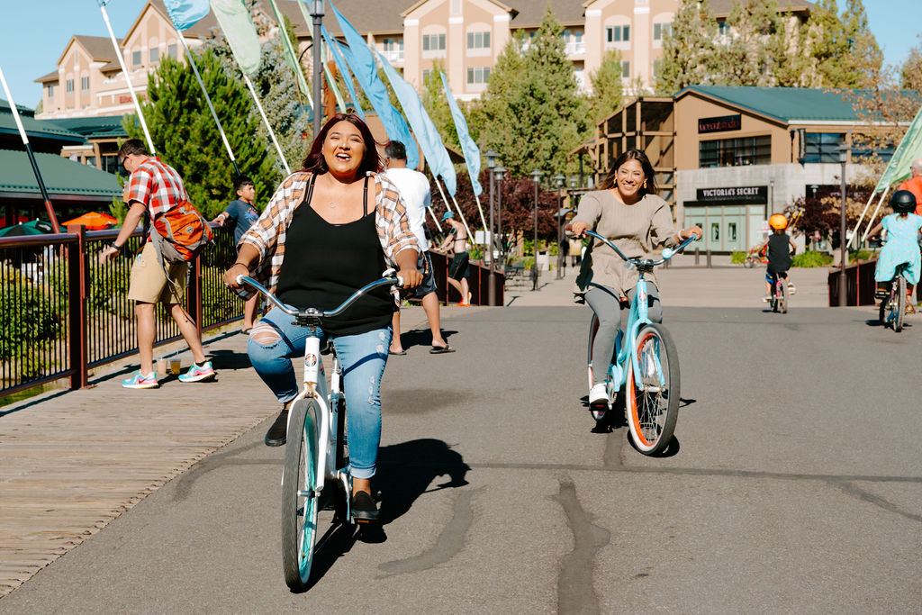 Gal with brown and pink hair riding her bike over the flag bridge in the Old Mill District in Bend, Oregon laughing with her friend on a baby blue bike beside her.