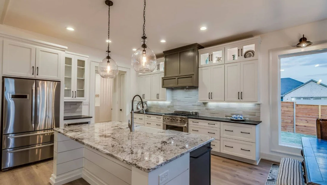 Modern kitchen featuring a large granite island with a sink, two hanging glass pendant lights, white cabinets, stainless steel refrigerator, and black countertops.