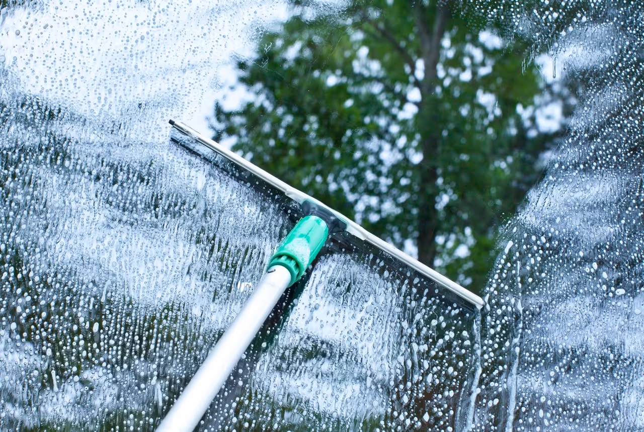 Squeegee cleaning a glass window covered with water droplets and soap suds with green trees blurred in the background.