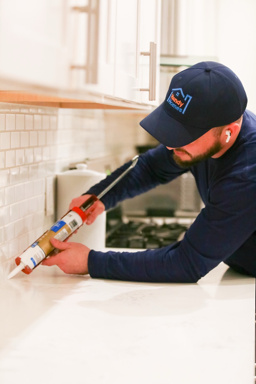 Man wearing a Handy Homes cap applying sealant along a kitchen countertop backsplash.