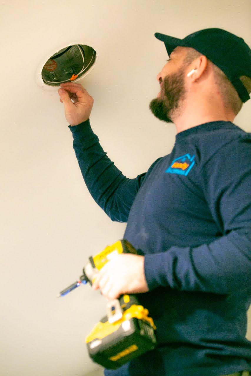 Man in a black cap and navy shirt working on an electrical fixture in the ceiling holding a yellow cordless drill.