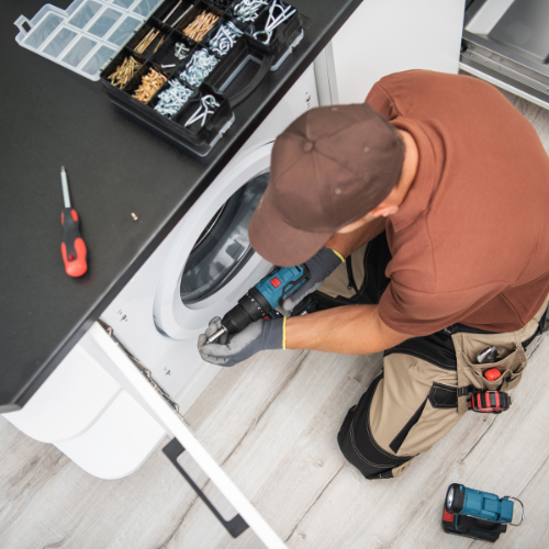 Technician wearing gloves and brown cap repairing a front-loading washing machine with a cordless drill.