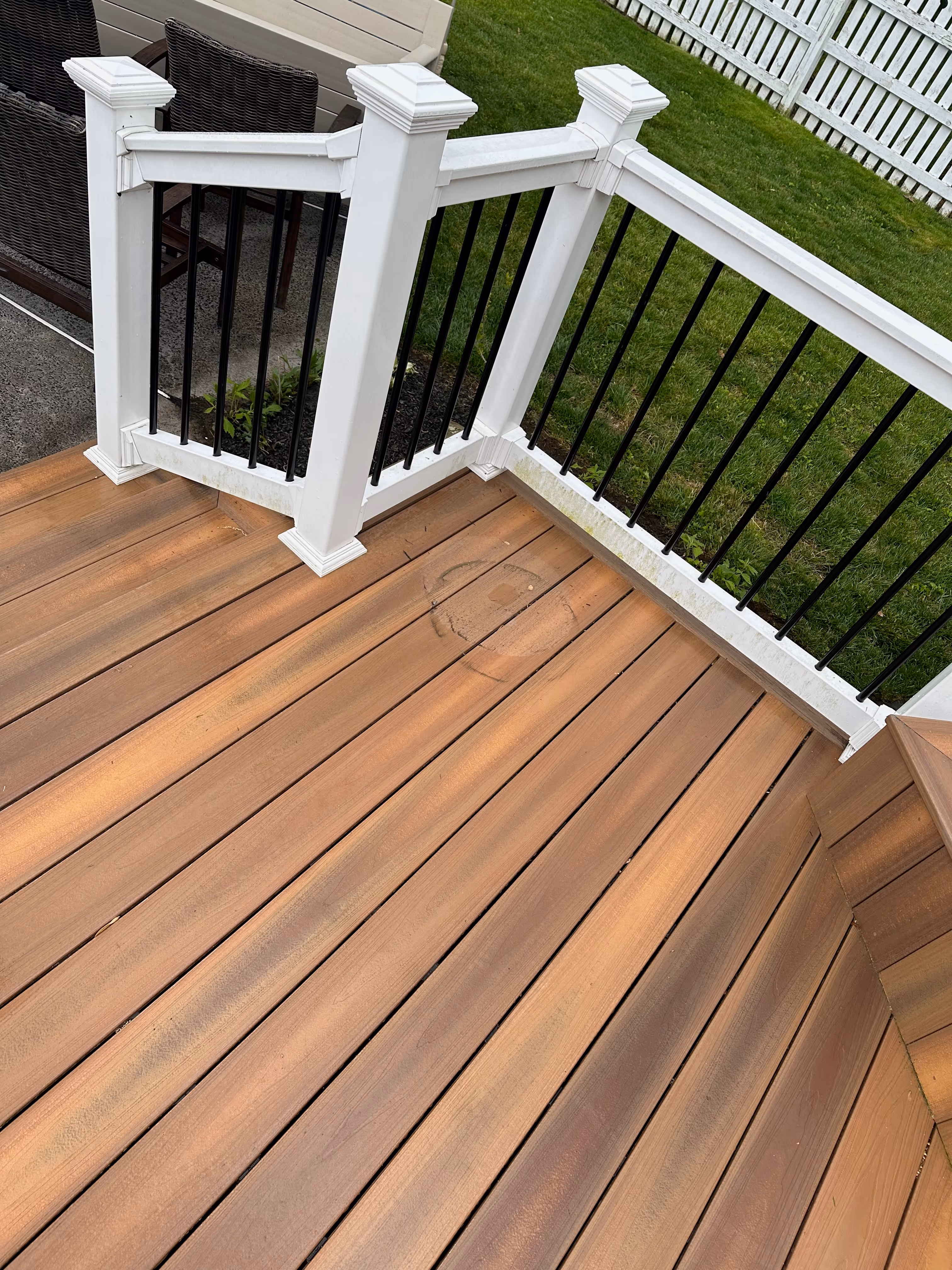 Wooden deck corner with white railing posts and black balusters next to green grass and a white fence.