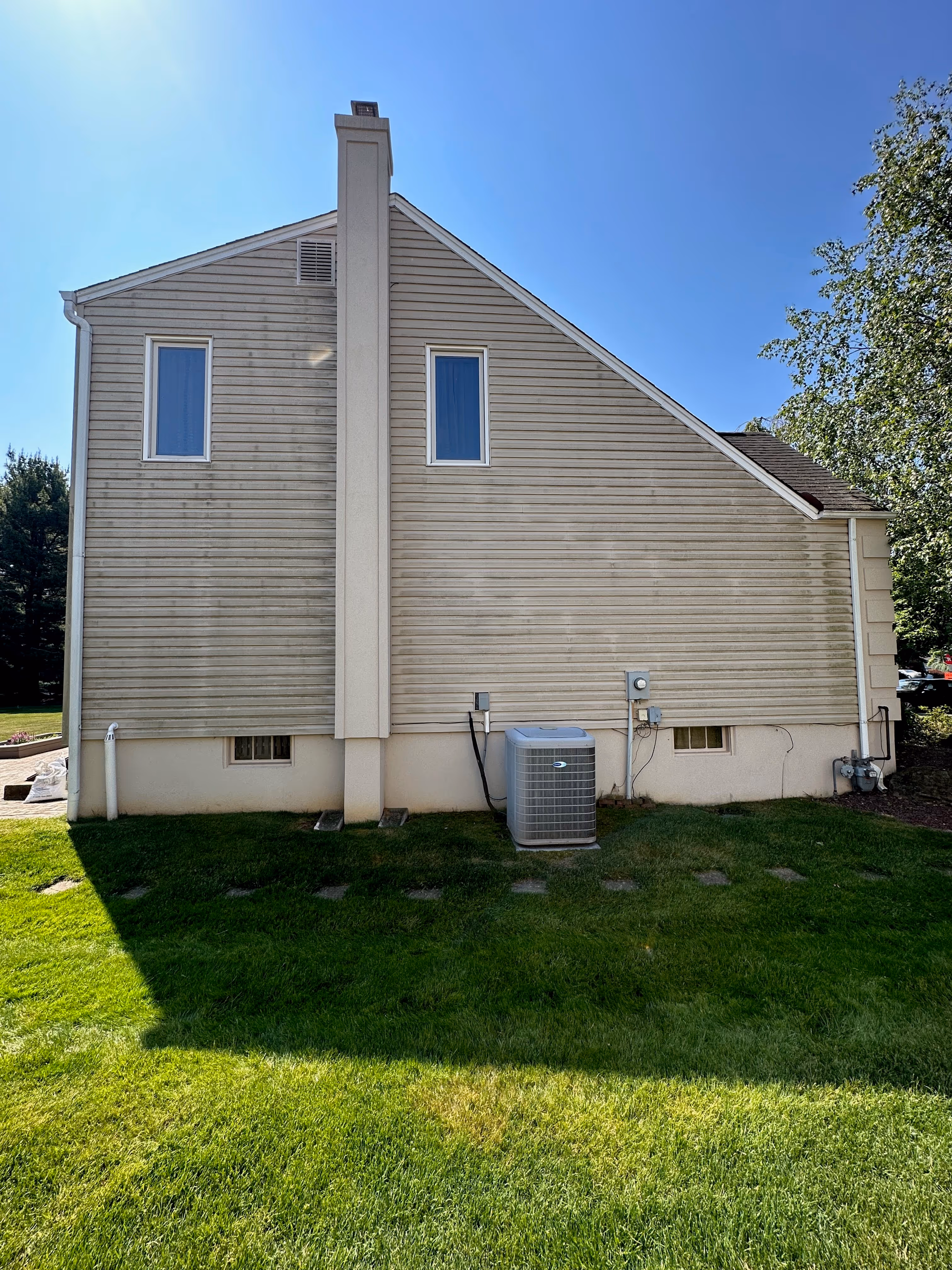 Side view of a beige house with two windows, a chimney, and an air conditioning unit on the green lawn under a clear blue sky.