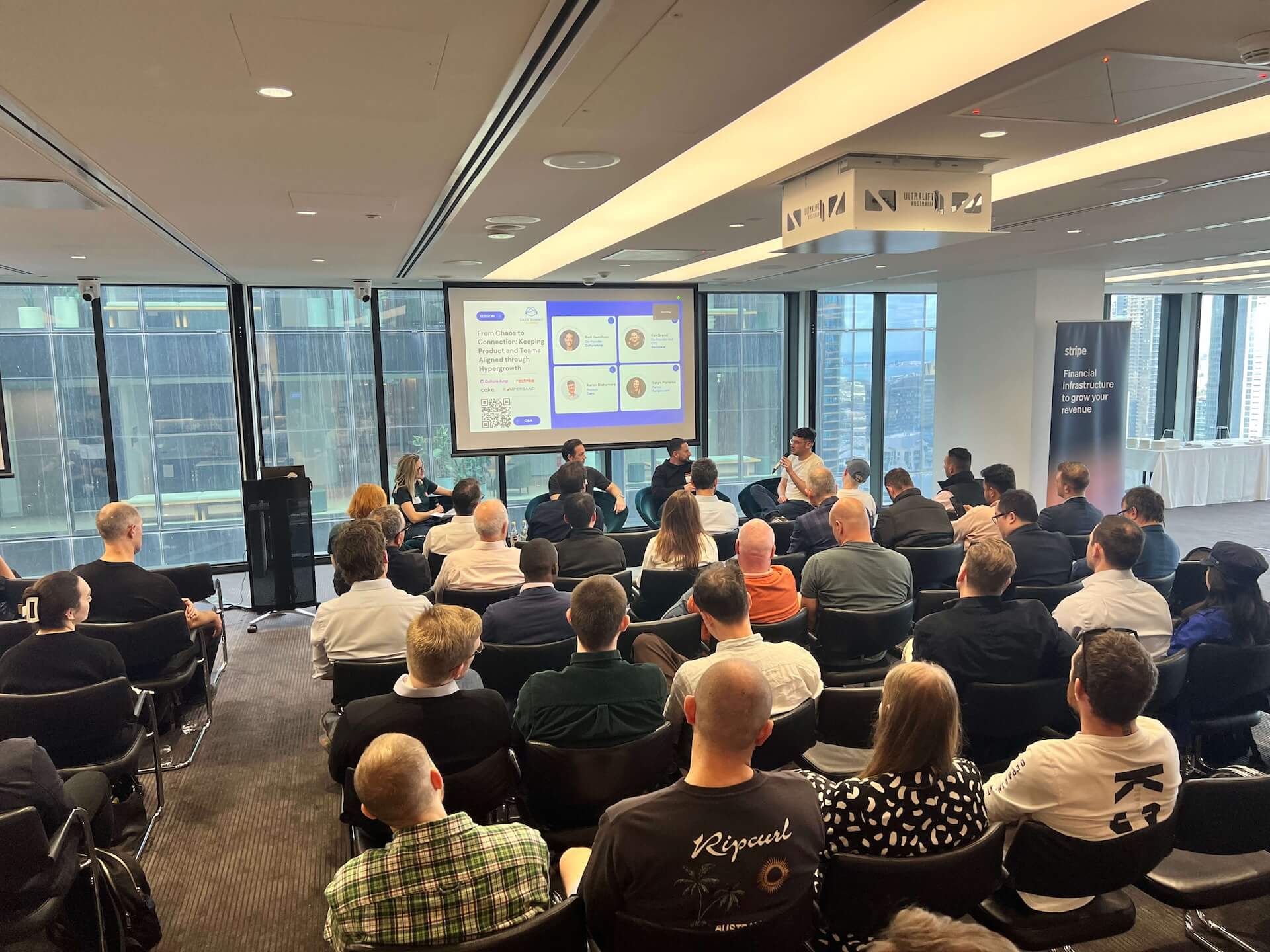 Audience attending a panel discussion in a modern conference room with large windows and a city view.