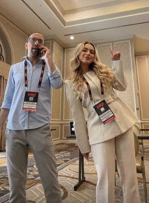 Man with glasses on phone and woman in beige suit flashing peace sign in conference room.